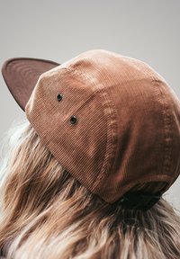 Braune Cord-Umhütze mit einem dunkelbraunen Schirm, ausgestattet mit zwei schwarzen Belüftungsöffnungen und einem schwarzen verstellbaren Riemen auf der Rückseite.