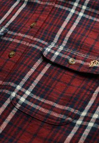 Camisa de franela a cuadros roja y azul marino con acentos en blanco, que presenta un diseño de botones, bolsillo y detalles de hardware de latón visibles.