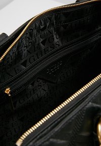 Black handbag interior with zippered pocket; textured lining features repeating "GUESS" pattern; gold hardware accents visible.