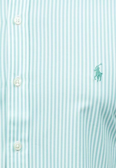 Gombos, világoskék és fehér függőleges csíkos ing, jobb oldalon egy kis hímzett logóval, amelyen egy lovon ülő pólójátékos látható.