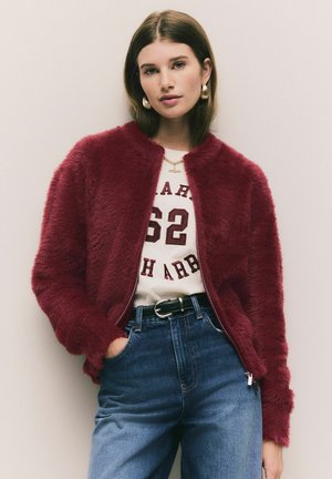 Červená huňatá bomber bunda so zipsom, nosená cez biele tričko s vínovými nápismi. V kombinácii s modrými vysokými džínami a čiernym opaskom.