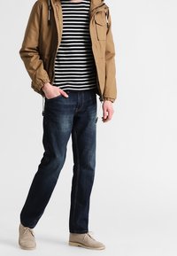 Chaqueta con capucha marrón sobre una camiseta a rayas negra y blanca, combinada con jeans de un azul oscuro y zapatos de un beige claro. Estilo casual, con un corte relajado.