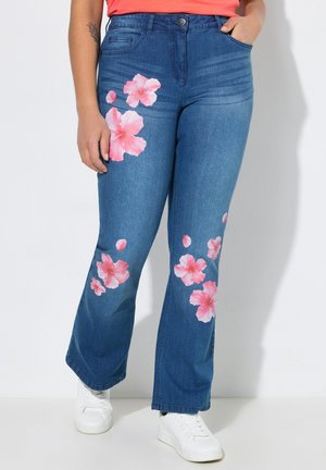 Blauwe flare jeans met roze bloemenprints, gecombineerd met witte sneakers, één hand in de zak en een tatoeage zichtbaar op de arm.