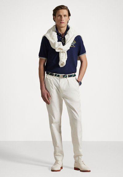 Navy polo shirt with white trims, cream trousers, white knitted sweater tied around neck, green and patterned belt, and light shoes.