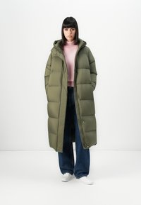 Kvinde står i en lang olivengrøn puffjakke, lyserød rullekrave, brede mørke jeans og hvide sneakers foran en ensfarvet hvid baggrund.