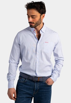 Man draagt een lichtblauw-wit gestreept overhemd met knopen en een borstzak, gecombineerd met een donkerblauwe spijkerbroek, heeft zijn hand in zijn zak en kijkt naar beneden.
