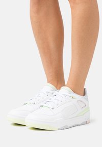 Chaussures de sport blanches avec une tige en cuir lisse, des accents vert clair, une languette rembourrée et une semelle en caoutchouc texturée présentant un design minimaliste.