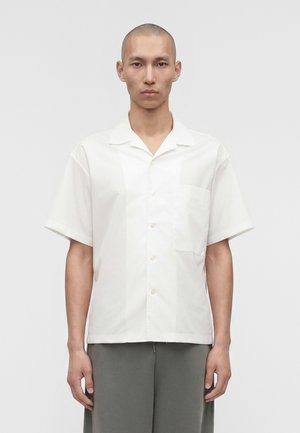 Άντρας με ξυρισμένο κεφάλι, που φοράει λευκό πουκάμισο με κοντά μανίκια και γκρι παντελόνι, στέκεται μπροστά σε μία απλή γκρις φόντο.