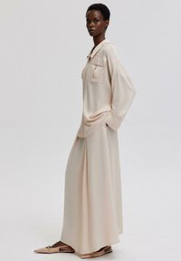 Ljust beige, långärmad blus med två bröstfickor, matchad med en flödande kjol. Modellen bär spetsiga platta skor. Slät textur.