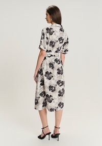 Sukienka do kolan w kremowym kolorze z czarnymi kwiatami, z krótkimi rękawami, kołnierzykiem i paskiem w talii. Noszona z czarnymi sandałami na obcasie.