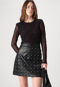 Svart mesh langermet topp med røde hjerte mønstre, kombinert med et svart lær miniskjørt pyntet med sølv-nagler. Modellen holder en vevd veske.