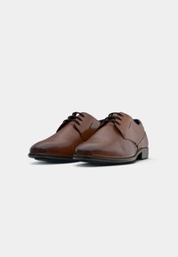 Scarpe da dress in pelle marrone da uomo, con lacci neri e tacchi bassi, esposte su uno sfondo bianco semplice.