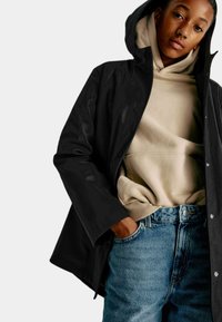 Sort vandtæt jakke med hætte, knaplukning, båret over en beige hoodie og lyseblå jeans. Glat tekstur, afslappet design.