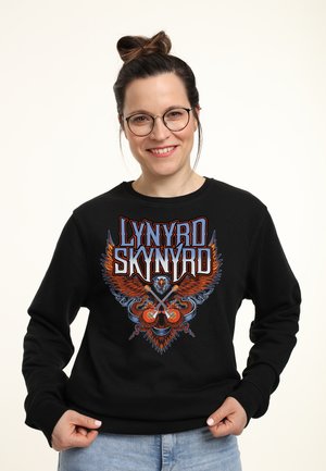 Smilende kvinde med briller iført sort Lynyrd Skynyrd sweatshirt med ørn og krydsede guitarer design.