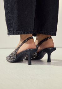 Crne salonke s trakicom oko pete, s teksturiranim, ukrašenim gornjim dijelom, šiljastim vrhom i tankom visokom petom. Potplat je gladak i blago sjajni.