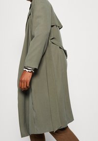 Długi, oliwkowo-zielony trencz o luźnym kroju, z wiązaniem z przodu i rozcięciami po bokach; wykonany z miękkiej, lekkiej tkaniny.
