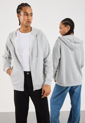 To personer med fletninger iført lysegrå hættejakker med lynlås, den ene vender fremad med en hvid skjorte og sorte bukser, den anden vender bort i blå jeans.