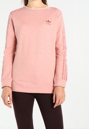 Kvinde iført en pink Adidas sweatshirt med lange ærmer og logo på brystet, kombineret med mørke bukser, stående foran en ensfarvet baggrund.