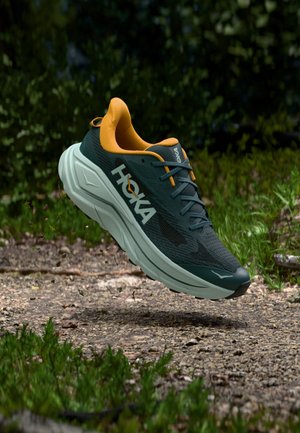 HOKA Laufschuh mit dunkelgrünem Stoffobermaterial, orangefarbenen Akzenten, dicker weißer Zwischensohle und strukturiertem Profil an der Außensohle, in der Luft schwebend dargestellt.