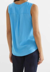 Ljusblå ärmlös topp med rundad halsringning, slät textur och en plissédetalj på baksidan, bärs med mörka byxor.