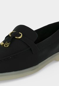 Mocassin noir à enfiler avec semelle blanche, orné d'un anneau décoratif doré et d'un pompon sur le dessus à l'avant.