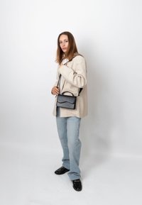 Jeune femme portant un blazer beige, un jean bleu clair et des mocassins noirs, tenant un sac bandoulière noir sur un fond blanc uni.