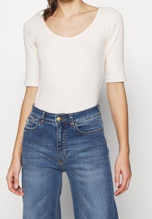 Kobieta nosząca kremową bluzkę z okrągłym dekoltem i półdługim rękawem, włożoną w wysokiej talii niebieskie dżinsy z denimu, stojąca na jednolitym tle.