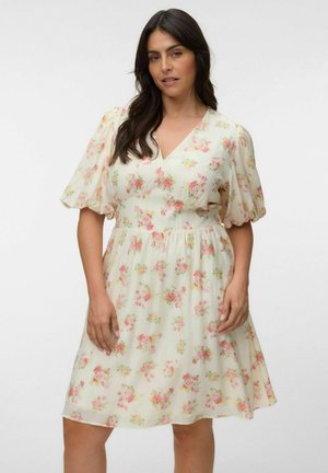 Frau trägt ein knielanges, hellcremefarbenes Kleid mit pinkem Blumenmuster und kurzen Puffärmeln, steht vor einem schlichten weißen Hintergrund.