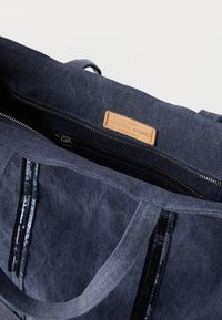 Τσάντα tote από ύφασμα σε ναυτικό μπλε με δερμάτινη ετικέτα· διαθέτει διαμέρισμα με φερμουάρ και γυαλιστερές παγιέτες κατά μήκος των πλευρών. Ομαλή υφή.