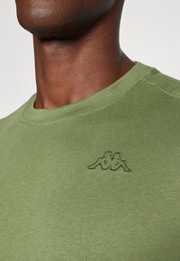 Ελιά πράσινη βαμβακερή μπλούζα με στρογγυλή λαιμόκοψη, που διαθέτει ένα διακριτικό υφασμένο λογότυπο στο στήθος και απαλό υφή.