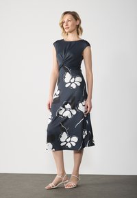 Marinefarbnes Midikleid mit kurzen Ärmeln, gedrehtem Oberteil und weißem Blumenmuster. Kombiniert mit hellen, sandalenartigen Schuhen. Glatte Stofftextur.