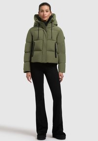 Giacca puffer verde oliva con cappuccio, caratterizzata da un taglio boxy, due tasche nere con zip e lacci neri per le regolazioni.