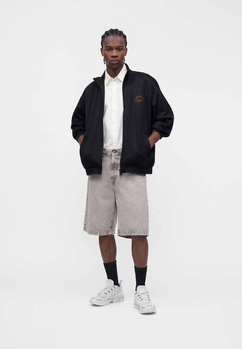 Mann mit geflochtenem Haar trägt schwarze Reißverschlussjacke, weißes Hemd mit Knöpfen, graue knielange Shorts, schwarze Socken und weiße Sneakers, steht frontal.