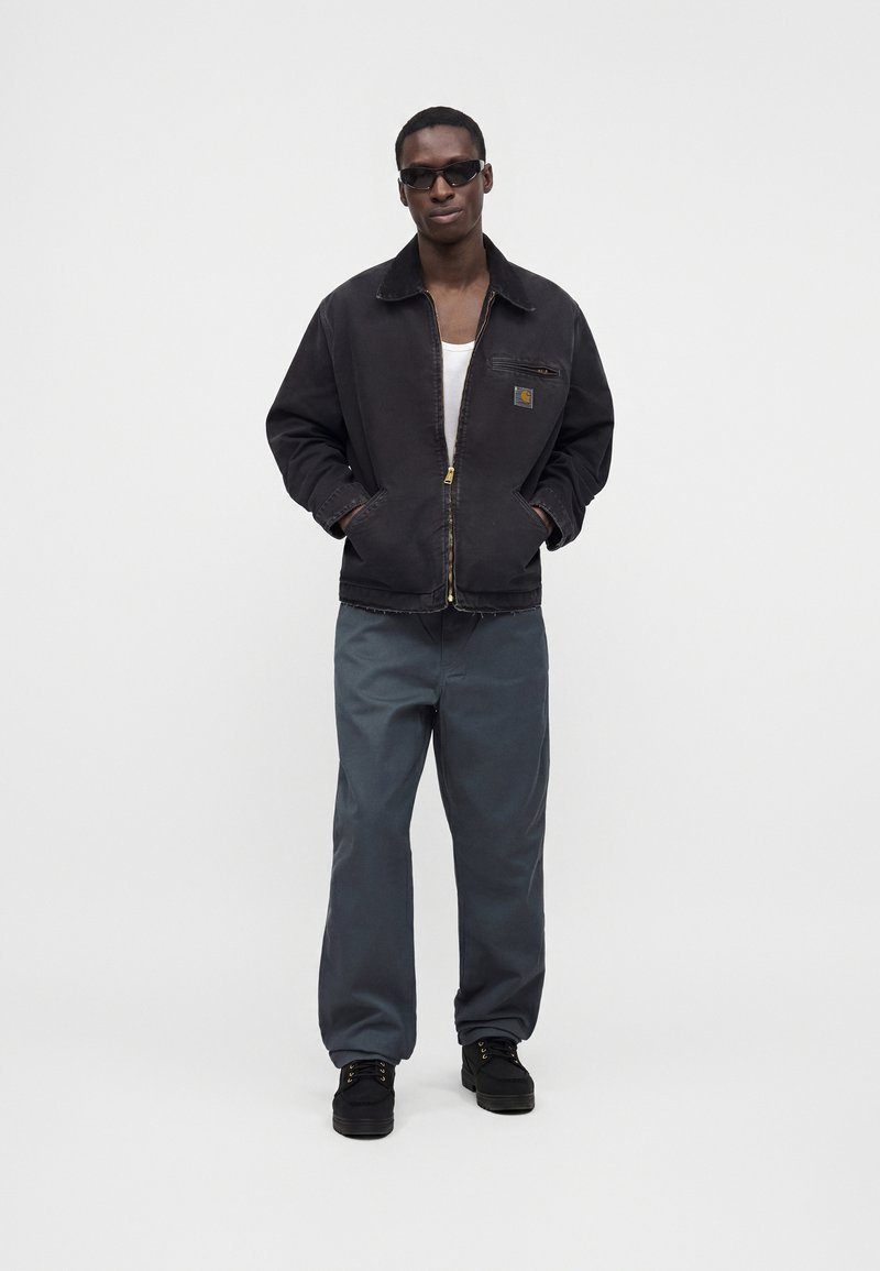 Homme portant une veste noire, une chemise blanche, un pantalon gris, des chaussures noires et des lunettes de soleil noires, debout les mains dans les poches contre un fond uni.
