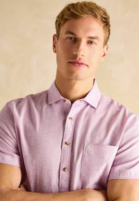 Heled lilla lühikeste varrukatega särk, mis on valmistatud tekstuursest kangast, esiküljel nööpidega, nähtava tasku ja beežide nööpidega.