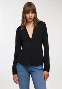 Donna con capelli lunghi fino alle spalle, che indossa una blusa nera con bottoni e ampi polsini, abbinata a jeans blu, in piedi di fronte a uno sfondo semplice.