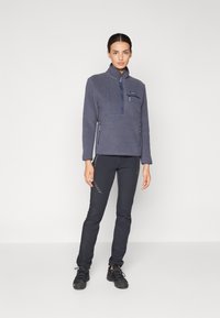 Felpa in pile grigio scuro con design a mezza zip, dotata di tasca sul petto e tasche laterali con zip, abbinata a pantaloni neri a sigaretta.