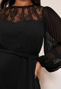 Fekete ruha csipke felsőrésszel, magas nyakkivágással, áttetsző redőzött hosszú ujjakkal és megkötős derékrésszel a testhez simuló sziluettért.