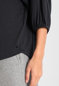 Černý, texturovaný top volného střihu s nabranými rukávy a zaobleným lemem, doplněný šedými, měkkými kalhotami podobnými fleecovým.