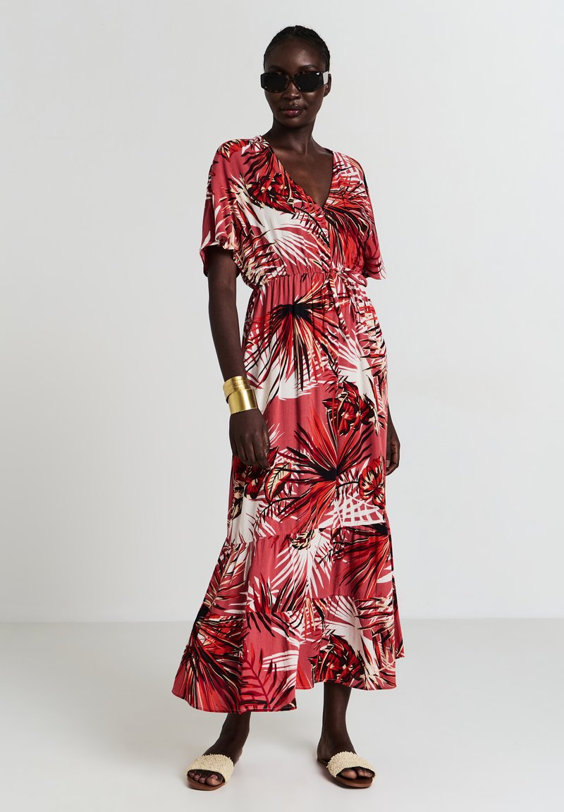 Robe maxi d'un rouge vibrant avec un motif de feuilles tropicales, taille élastique, manches courtes et silhouette fluide, assortie à des sandales neutres.