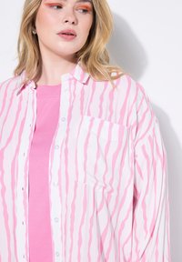 Leichtes, durchsichtiges, kurzärmeliges Hemd mit rosa und weißen wellenförmigen Streifen, versehen mit einer Brusttasche und über einem einfarbigen rosa Oberteil getragen.