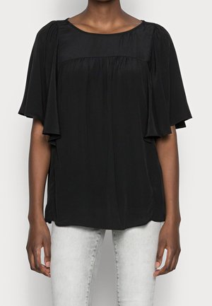 Kvinde iført en løs sort bluse med vide ærmer og rynkede detaljer, kombineret med lysegrå jeans.