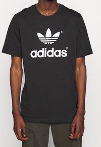 Černé bavlněné tričko s velkým bílým logem Adidas trefoil. Krátké rukávy a klasický kulatý výstřih. Měkká textura.