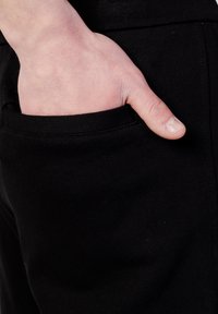 Pantaloni neri con una tasca laterale. Una mano è appoggiata nella tasca, mostrando la tessitura liscia e la vestibilità del tessuto.