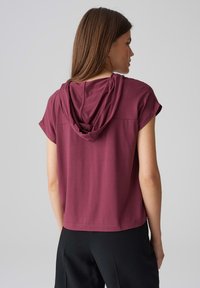 Top corto bordeaux a maniche corte con cappuccio, caratterizzato da una vestibilità rilassata e una texture liscia, abbinato a pantaloni neri.