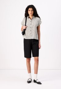 Camicia a maniche corte nera con motivo crema abbinata a pantaloni corti neri su misura. Il modello indossa calze bianche e ballerine nere, mentre tiene una borsa piccola.