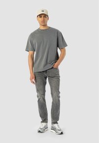 Joven con una camiseta gris, jeans grises desgastados, zapatillas plateadas y una gorra beige, de pie con una mano en el bolsillo.
