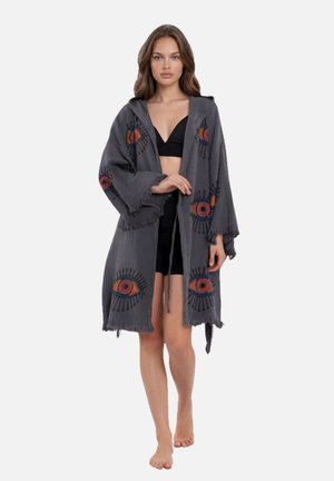 Vrouw die een lange grijze badjas draagt met kleurrijke oogpatronen, een zwart cropped topje en shorts, blootsvoets staand tegen een effen achtergrond.