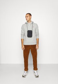 Grijze hoodie met trekkoorden, een klein logo, gecombineerd met bruine corduroy broek. Een zwarte pouch hangt om de nek, gedragen met witte sneakers.
