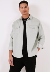 Camicia azzurrina chiaro con bottoni, realizzata in tessuto morbido, caratterizzata da due tasche frontali e un design delle maniche piegate. Indossata sopra una camicia nera.